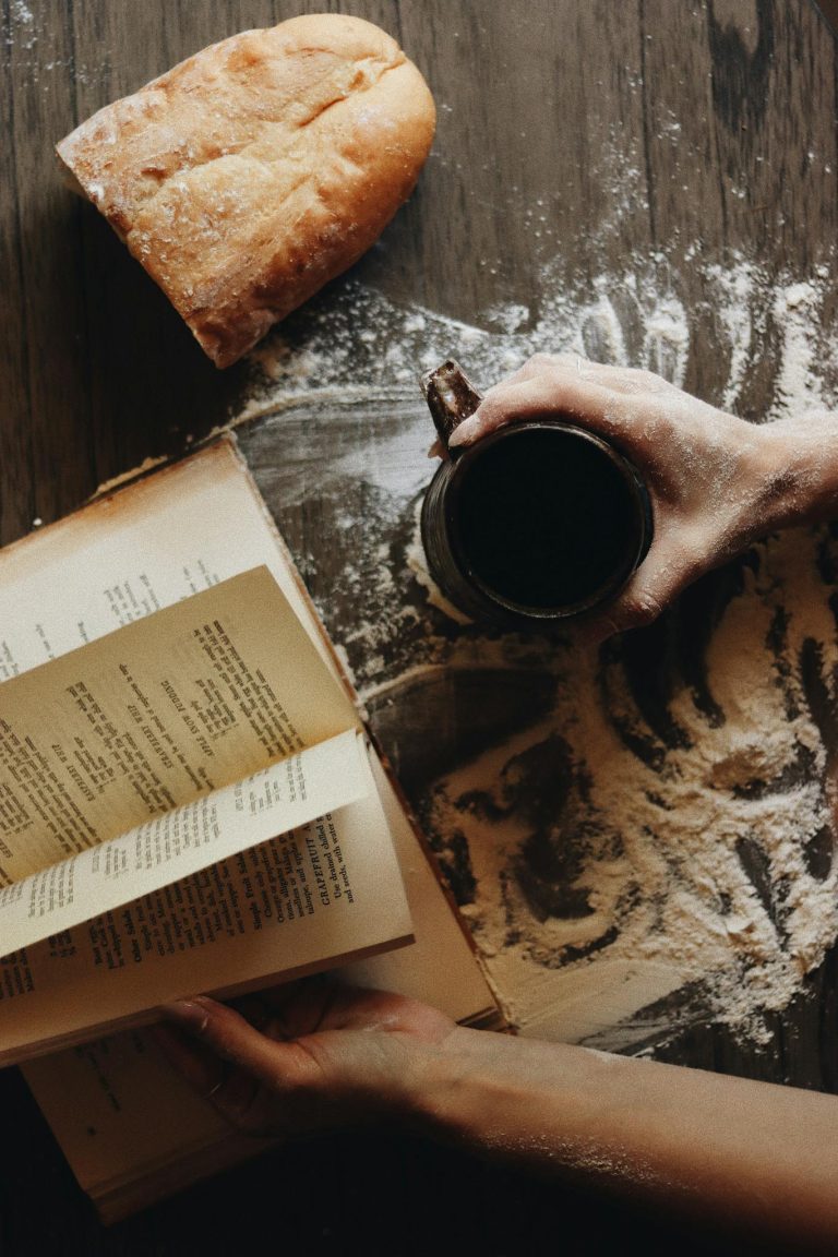 A cozy scene with a loaf of bread, recipe book, and a coffee mug on a flour-dusted table.