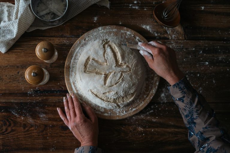 A person carving designs into bread dough on a rustic wood table, highlighting traditional baking.
