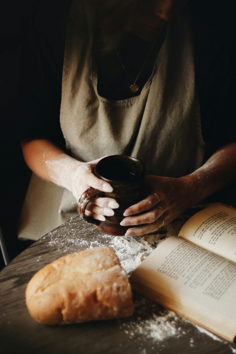 A warm, cozy kitchen scene featuring fresh bread, a recipe book, and a person enjoying coffee.